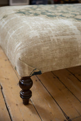 Close-up of antique rug ottoman corner; beige textile, visible foundation, and dark turned leg.