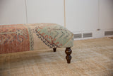 Vintage Oushak rug fragment ottoman side with distressed rust, mint geometric patterns, and dark wood leg.