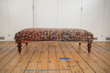 Vintage Persian rug ottoman, side view, with navy, coral, ivory pattern and dark turned legs.