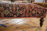 Detail of vintage Persian rug ottoman, navy, blush, orange tribal pattern, turned wooden leg.