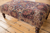 Antique Kerman rug fragment ottoman, patterned beige, red, blue upholstery showing wear.