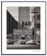 A black and white photograph of a downtown New York restaurant and bar with vintage cars on a cobblestone street.
