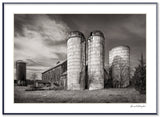 Black and white photograph of a rustic New York barn with weathered silos under a dramatic sky.