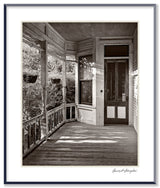 Black and white photograph of a weathered wooden side porch with sunlight creating strong shadows across the floor.