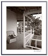 Black and white photograph of a rustic wooden side porch with a wicker chair and white railing.