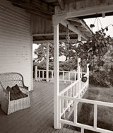 View along a rustic wooden side porch with a wicker chair, captured in a monochrome photograph.
