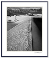 A black and white photograph of a sand dune, featuring a textured crest against a deep shadow and distant mountains.