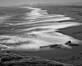 Long-exposure black and white photograph of soft waves on a dark, sandy beach.