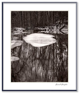Framed black and white photograph showing tree reflections surrounding a central patch of ice on a pond.