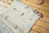 Corner of vintage Moroccan rug, shaggy ivory pile, dark playful motifs, and colorful fringe.