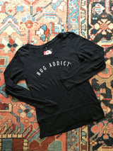 Black 'Rug Addict' long sleeve shirt with arched white block letters laid flat.