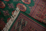 Vintage Bokhara carpet showing emerald green pile, rust gul medallions, ivory fringe, and geometric patterned edge.