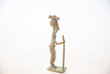 Profile view of a walking African copper figure with an oxidized patina, holding a staff.