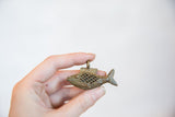 Hand holding the handmade African bronze fish pendant, showing its mesh body and aged texture.