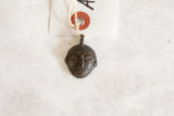 Vintage African bronze mask pendant laid flat, showing its tag and textured surface details.