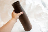 Hand holding the tall antique African vase, showing its dark wood patina and a visible split near the top.