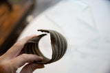 A hand holds the antique bronze cuff, showing its open shape and aged, ribbed texture.