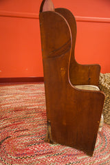 Antique wide plank settle chair's side, showing curved back, plaid seat, worn leg.