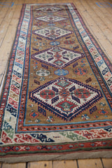 Angled view of an antique Caucasian runner showing repeating diamond medallions on a warm ochre field.