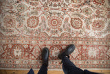 A person stands on a distressed vintage Meshed carpet, showing its worn floral pattern and texture.