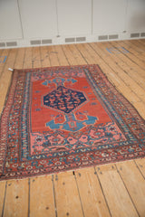 Vintage Kurdish Bijar rug at an angle, displaying a central navy diamond medallion on a cherry red field.