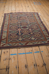 Angled view of a vintage Caucasian square rug with repeating octagonal guls in navy blue, red, and beige.