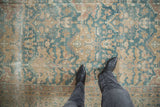 A person's feet on a distressed Agra carpet, revealing worn pile and dusty blue palmettes.