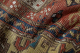 Visible hand-knotted weave texture with brick red, navy, sand geometric patterns on an antique Kazak rug.
