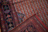 Antique Qashqai rug corner showcasing navy geometric vinery border, ruby red striped field, and fringed end.