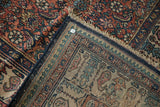 Folded corner of a vintage Persian rug showing the secured navy edge, fringe, and boteh border pattern.