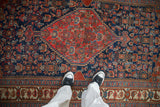 Feet in black sneakers on antique Bijar rug with indigo, cherry, teal Herati motif.