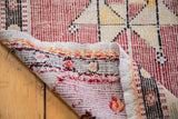 Distressed Jijim runner's folded corner, showing hand-woven texture, faded cranberry, and orange stitched edge.