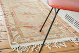 Corner of distressed Oushak rug with braided fringe, plain weave, and geometric red border.