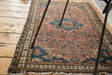 Distressed Malayer rug corner with worn dusty rose field, blue patterns, and tattered fringe.