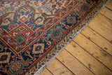 Corner view of distressed Heriz rug, showing brick red and navy geometric floral border with fringe.