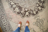 Distressed Oushak rug with feet, showing worn chocolate medallion, antique ivory, blush field.