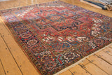 Vintage Heriz carpet corner showing cherry red field, navy blue scrolling floral border, and fringe.