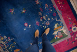 Person's feet on a blue Art Deco pictorial rug with floral tendrils and cochineal red border.
