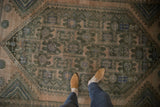 Distressed vintage rug with geometric floral arabesques and muted charcoal, dusty rose tones.