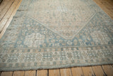 Angled view of a distressed Qashqai carpet's dusty blue corner spandrel and sand-colored field with floral motifs.