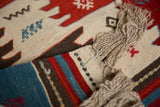 Folded edge of a Turkish Kilim rug showing natural ivory fringe and striped red, blue, charcoal bands.