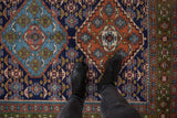 Feet on vintage Ardebil rug showing navy, terra cotta, green Herati-style medallion detail.