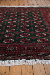 Angled view of a vintage Bokhara carpet highlighting the plush green pile and neat white fringe.