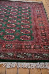 A vintage Bokhara carpet's corner, showing the plush green wool, detailed rust borders, and ivory fringe.
