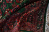 A folded corner showing the lustrous, plush pile of a Pakistani Bokhara carpet in rust, green, and ivory.