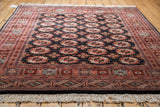 Angled view: Vintage Bokhara carpet, repeating gul medallions, charcoal field, terra cotta geometric borders.