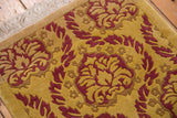 Corner view of embossed curvilinear Damask pattern in raspberry and saffron with ivory fringe.