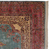 Corner of a vintage Kerman carpet showing the pink millefleur border, blue field, and clean cotton fringe.