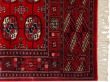 Corner of a vintage Bokhara rug showing a red gul medallion, intricate geometric borders, and full ivory fringe.