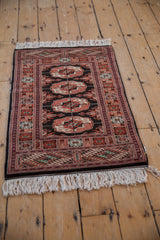 Angled view of a vintage Bokhara rug showing its repeating rust and ivory gul medallions on a black field.